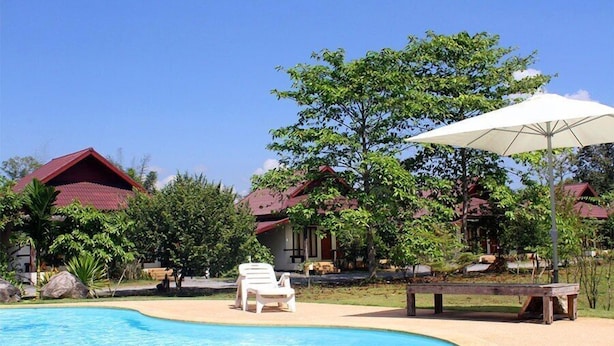 A pool area with lounge chairs and umbrellas, surrounded by trees and buildings with red roofs. - Bangyai Buri Resort (Surat Thani)