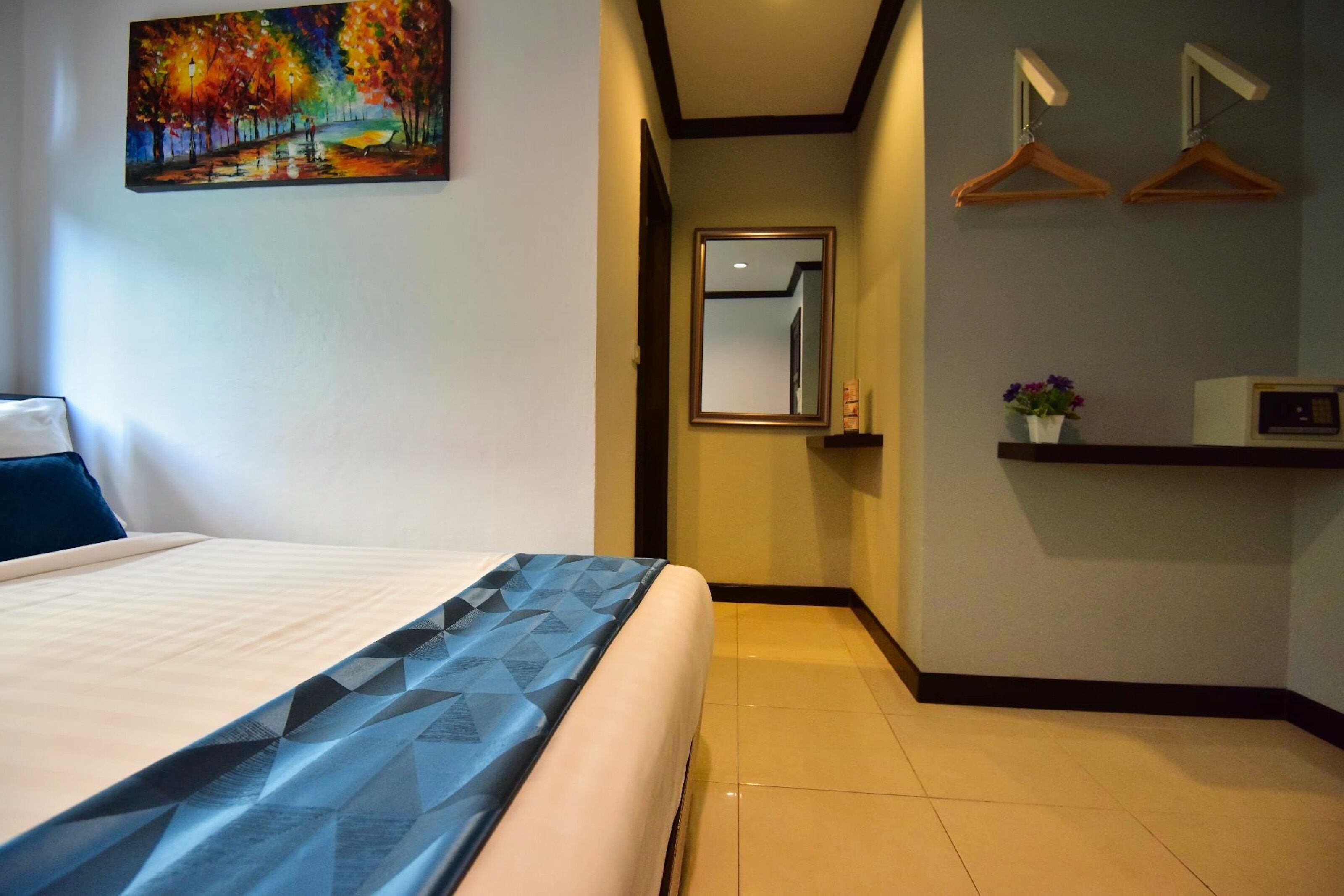 A hotel room with a bed, a painting on the wall, a mirror, and a shelf with a vase.