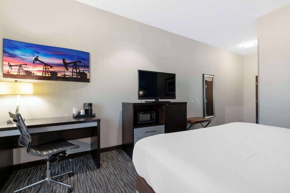 A hotel room with a bed, desk, chair, TV, and a painting of oil pumps.
