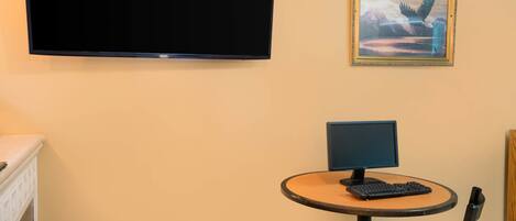 A room with a flat-screen TV mounted on the wall, a small round table with a computer and keyboard, a chair, and a framed picture above the table.