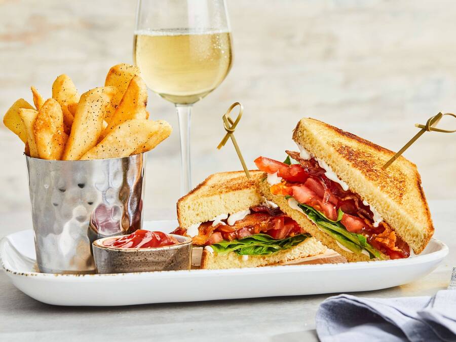 A plate with a grilled sandwich, a glass of white wine, and a container of fries.