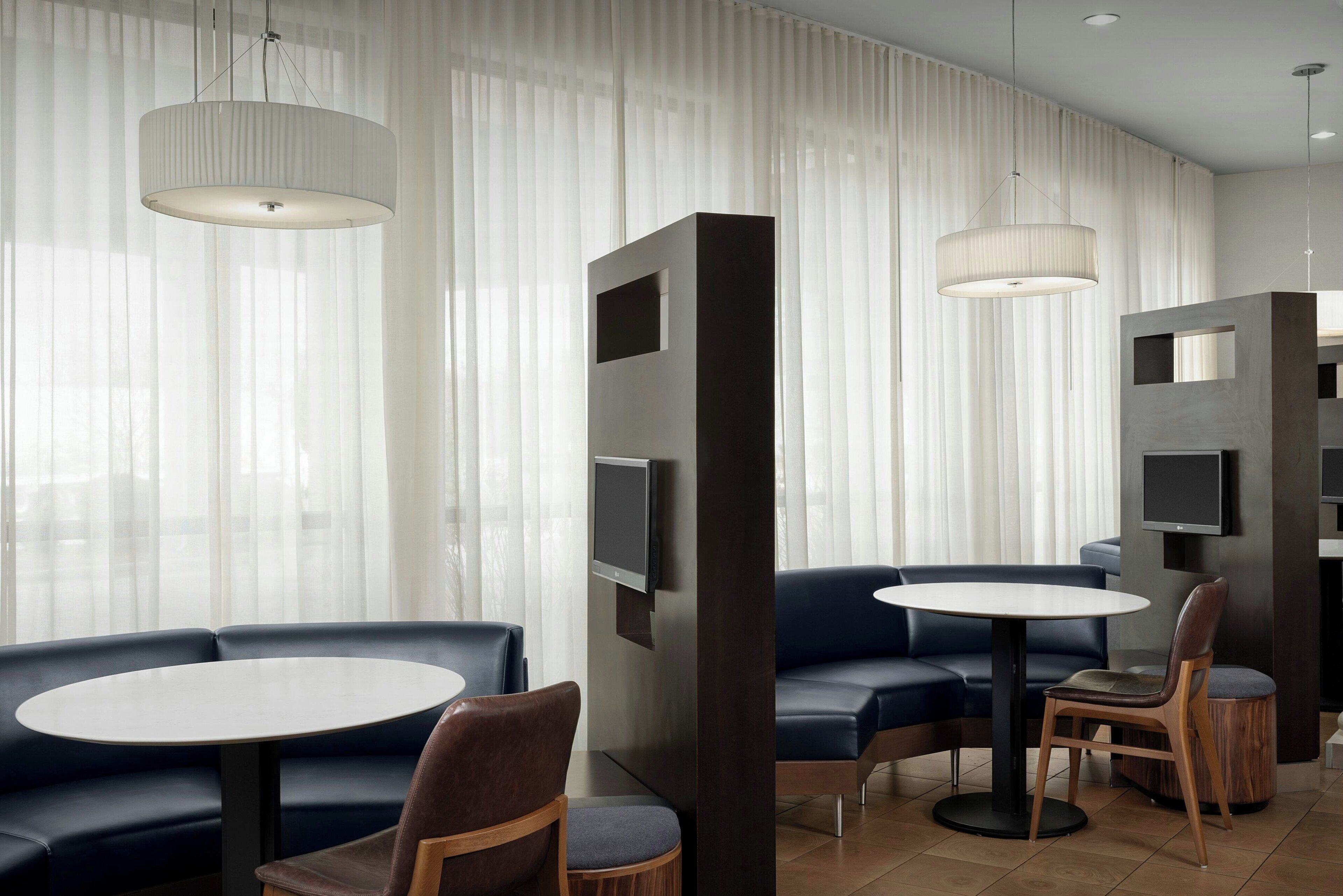 A modern hotel lobby with a round table, blue seating, and large windows with sheer curtains.