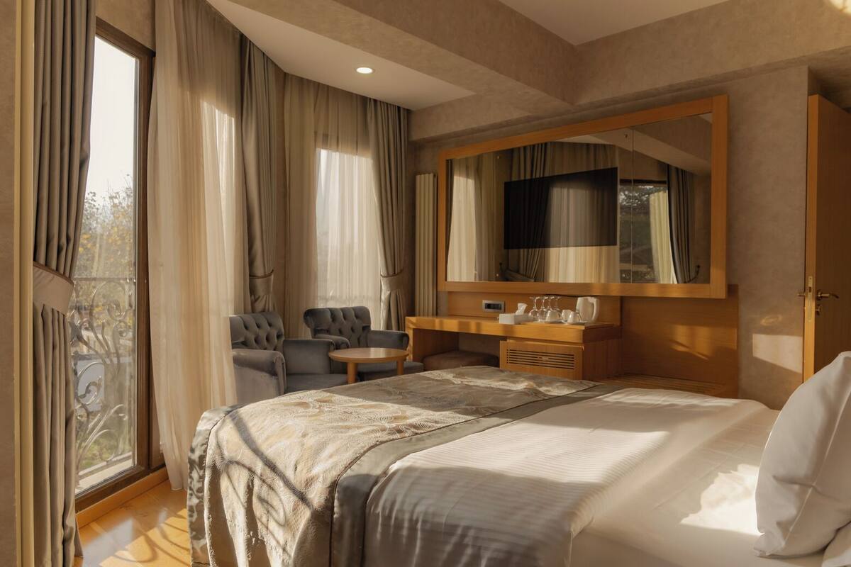 A hotel room with a large bed, a seating area with two chairs, a small table, a mirror, and a balcony with curtains.