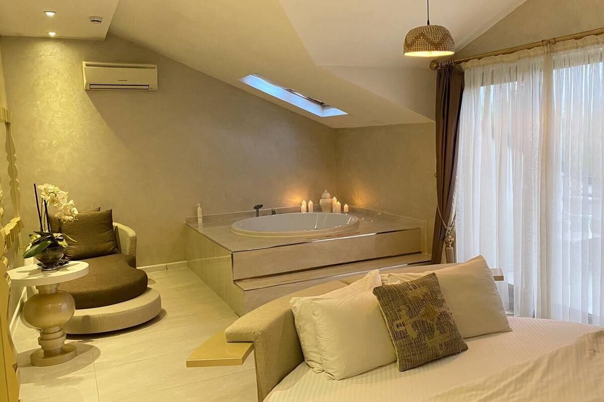 A modern bedroom with a large bed, a built-in bathtub, a round seating area, and a skylight.