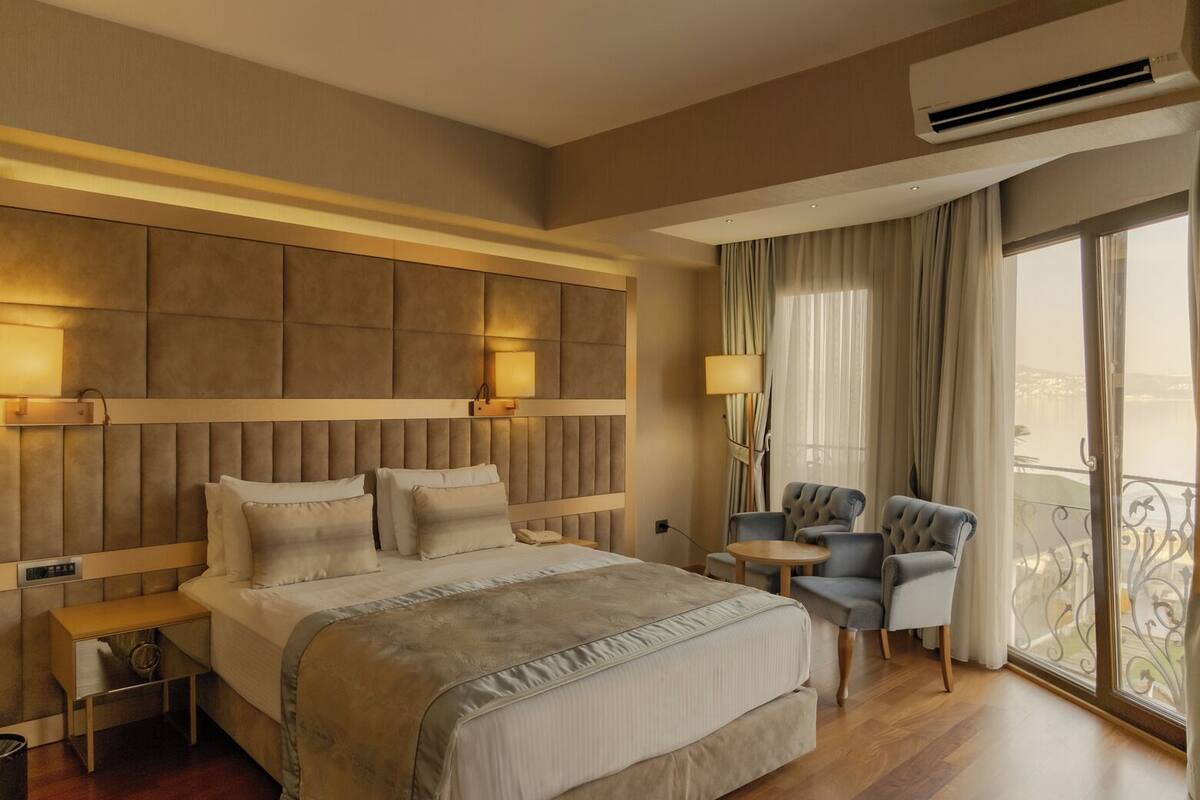 A hotel room with a large bed, bedside tables, a desk, two chairs, a balcony with a view, and an air conditioning unit.