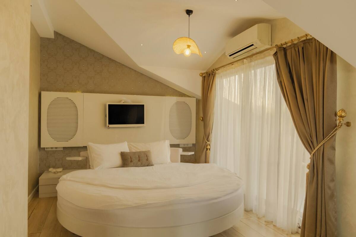 A bedroom with a large bed, a mounted TV, an air conditioning unit, and a window with curtains.