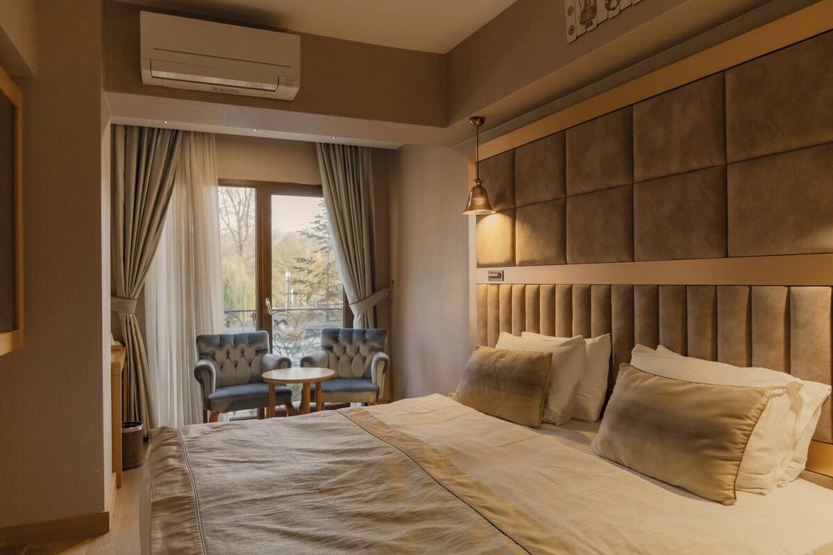 A hotel room with a large bed, a headboard with a padded design, a small round table, two armchairs, and a window with curtains.