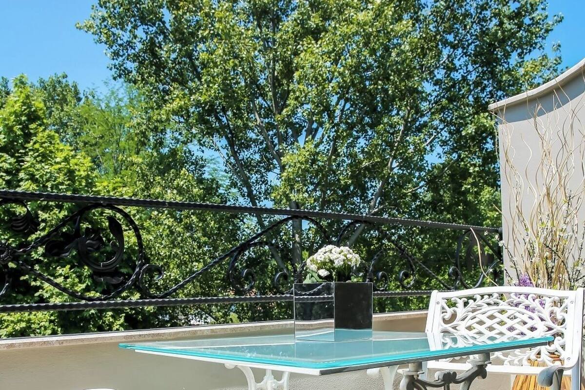 A patio with a glass table and white metal chairs, surrounded by trees and a white wall.