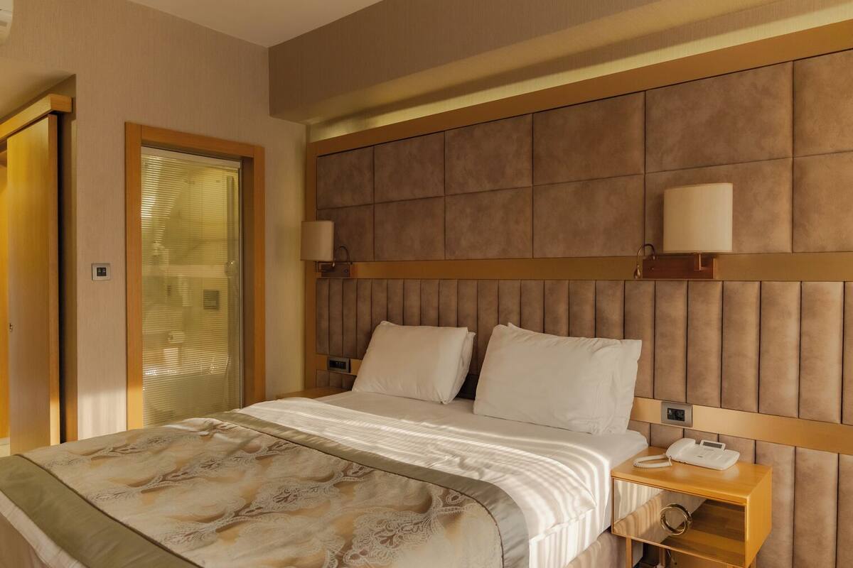 A hotel room with a bed, bedside tables, a wall-mounted lamp, and a door with blinds.