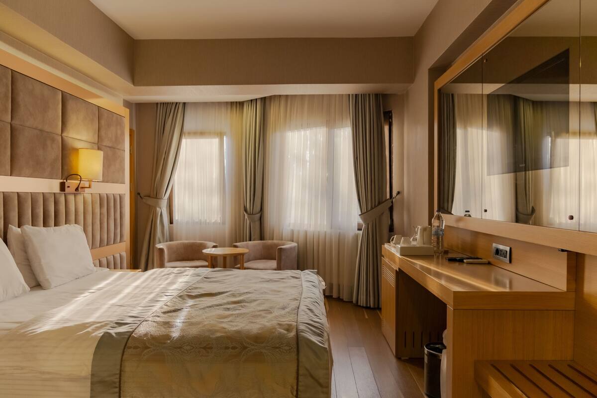 A hotel room with a large bed, a desk, a chair, a small table, a mirror, and curtains.