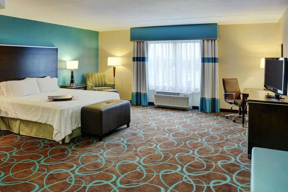 Ein Hotelzimmer mit Bett, Schreibtisch, Stuhl, Fernseher und einem Fenster mit Vorhängen.