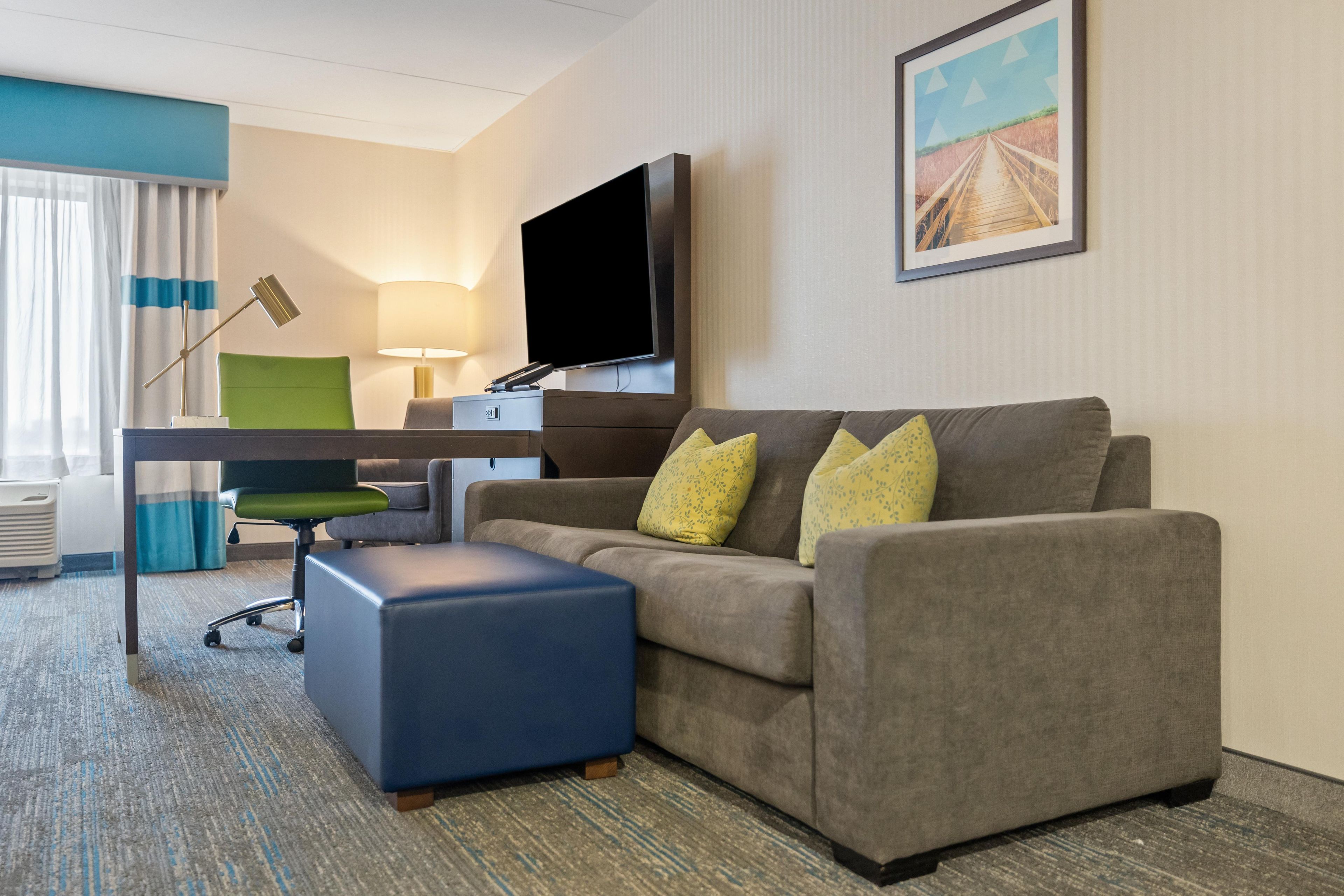 A hotel room with a sofa, a desk, a chair, a flat-screen TV, a lamp, and a painting on the wall.
