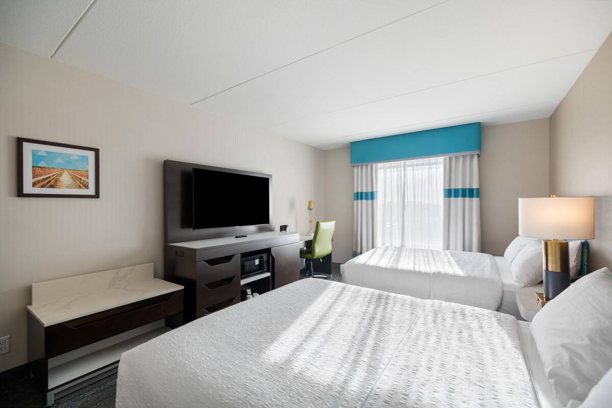 Ein Hotelzimmer mit Bett, Schreibtisch mit Flachbildfernseher, Stuhl, Nachttisch und einem Fenster mit Vorhängen.