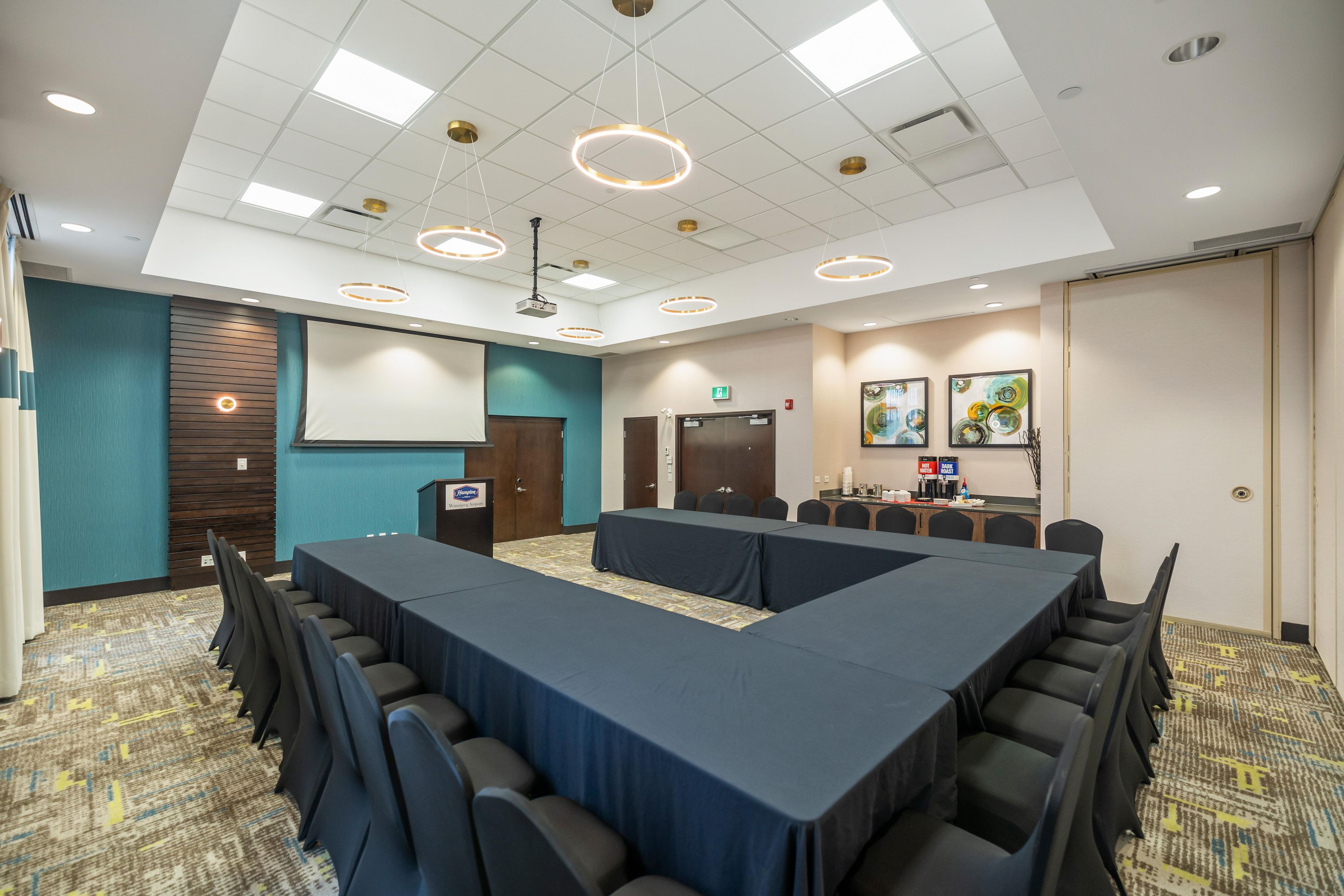 Un salón de conferencias con una mesa larga, sillas, un atril, una pantalla de proyector y una kitchenette con dispensadores de café y agua.