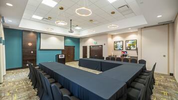 Un salón de conferencias con una mesa larga, sillas, un atril, una pantalla de proyector y una kitchenette con dispensadores de café y agua.