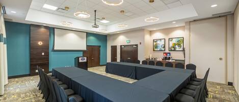 A conference room with a long table, chairs, a podium, a projector screen, and a kitchenette with coffee and water dispensers.