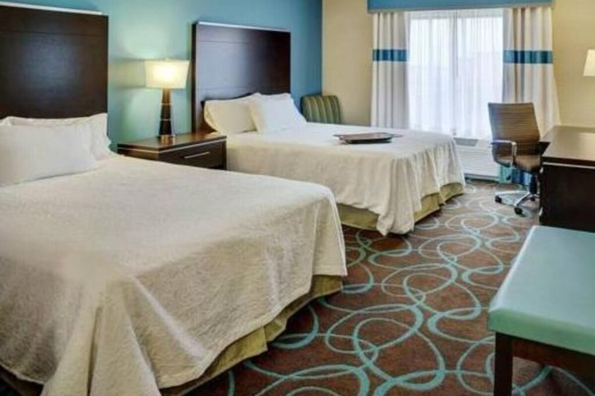 Une chambre d’hôtel avec deux lits, un bureau, une chaise et une fenêtre avec des rideaux.