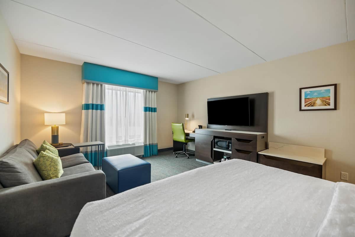 Ein Hotelzimmer mit einem Bett, einem Schreibtisch, einem Fernseher, einem Sessel und einem Fenster mit Vorhängen.