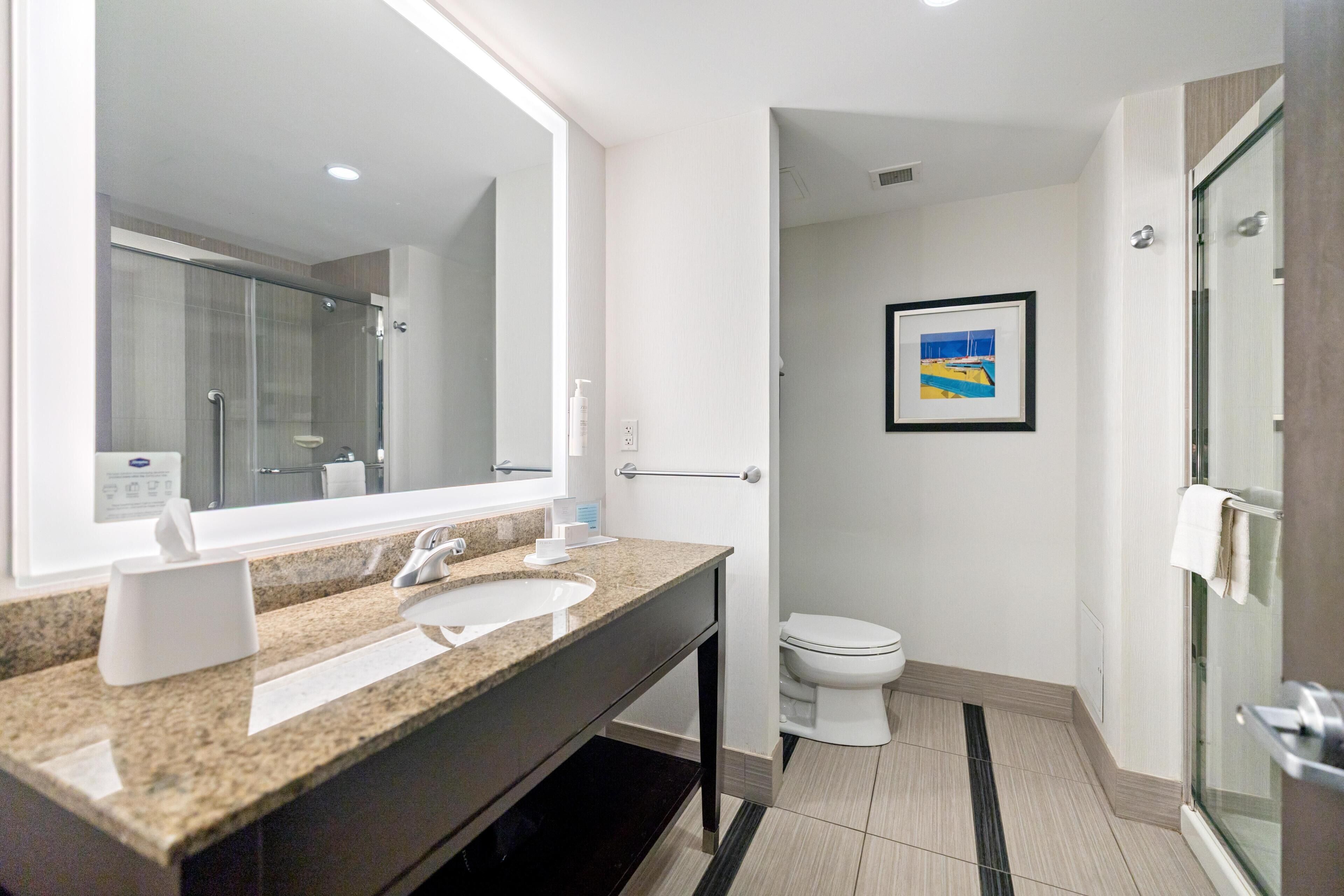 Un baño moderno con encimera de granito, un espejo grande, inodoro y cabina de ducha con mampara de vidrio.