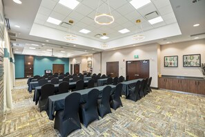 Une salle de conférence avec des rangées de tables et de chaises, un coin cuisine et un espace d’accueil.