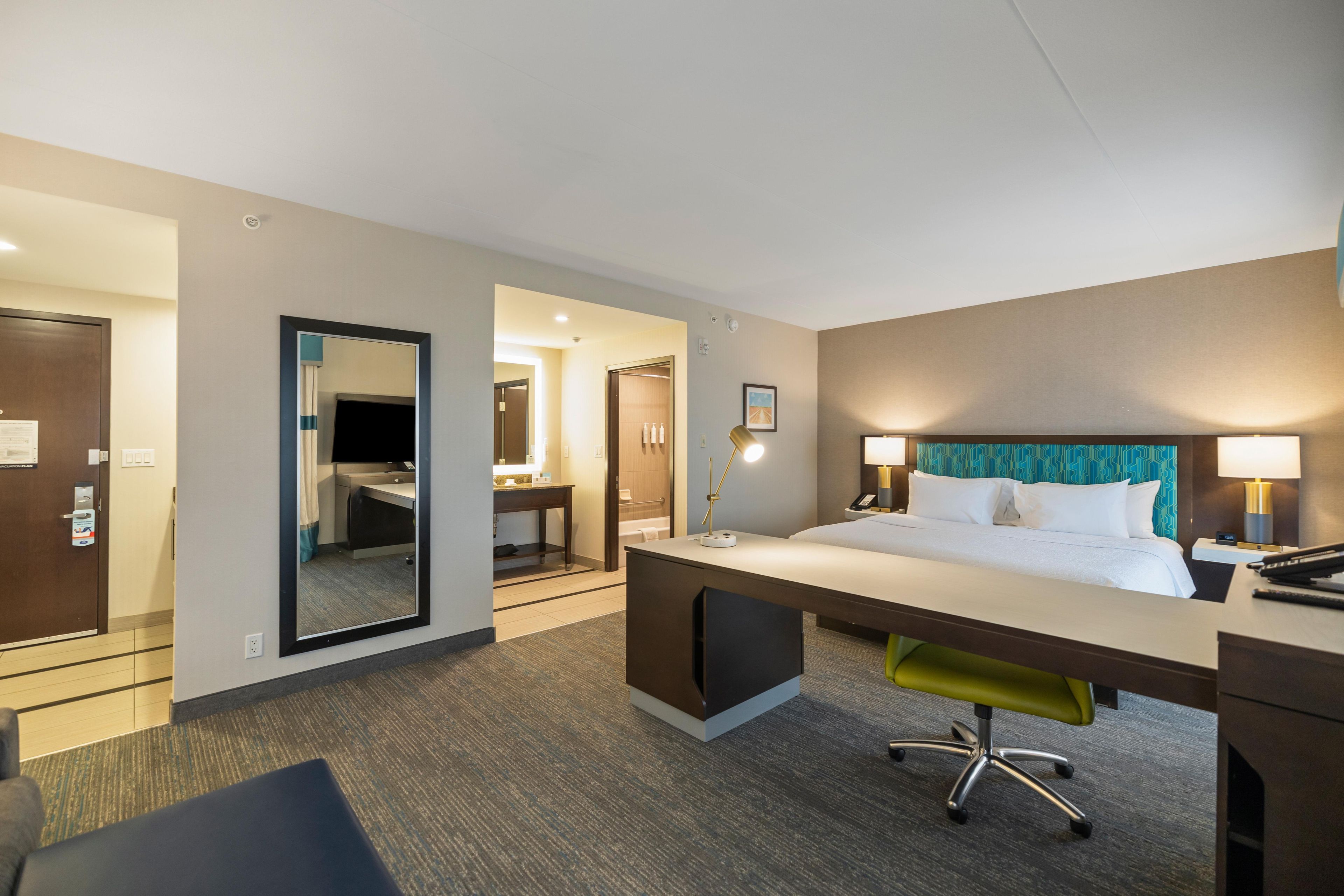 Habitación de hotel con cama, escritorio, silla y baño visible a través de una puerta abierta.