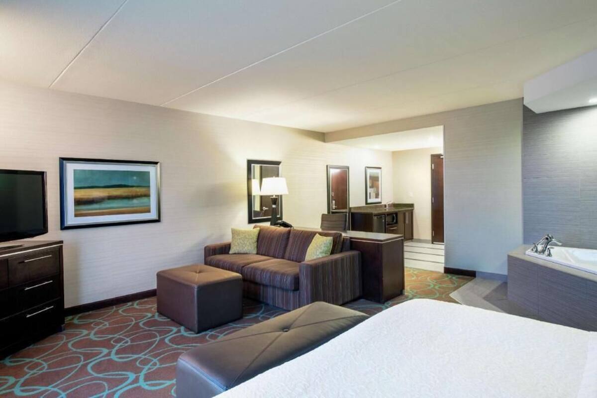 A hotel room with a bed, a sofa, a TV, a bathroom, and a painting on the wall.