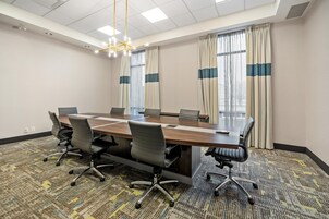 Un salón de conferencias con una larga mesa, sillas, una araña y ventanas grandes con cortinas.