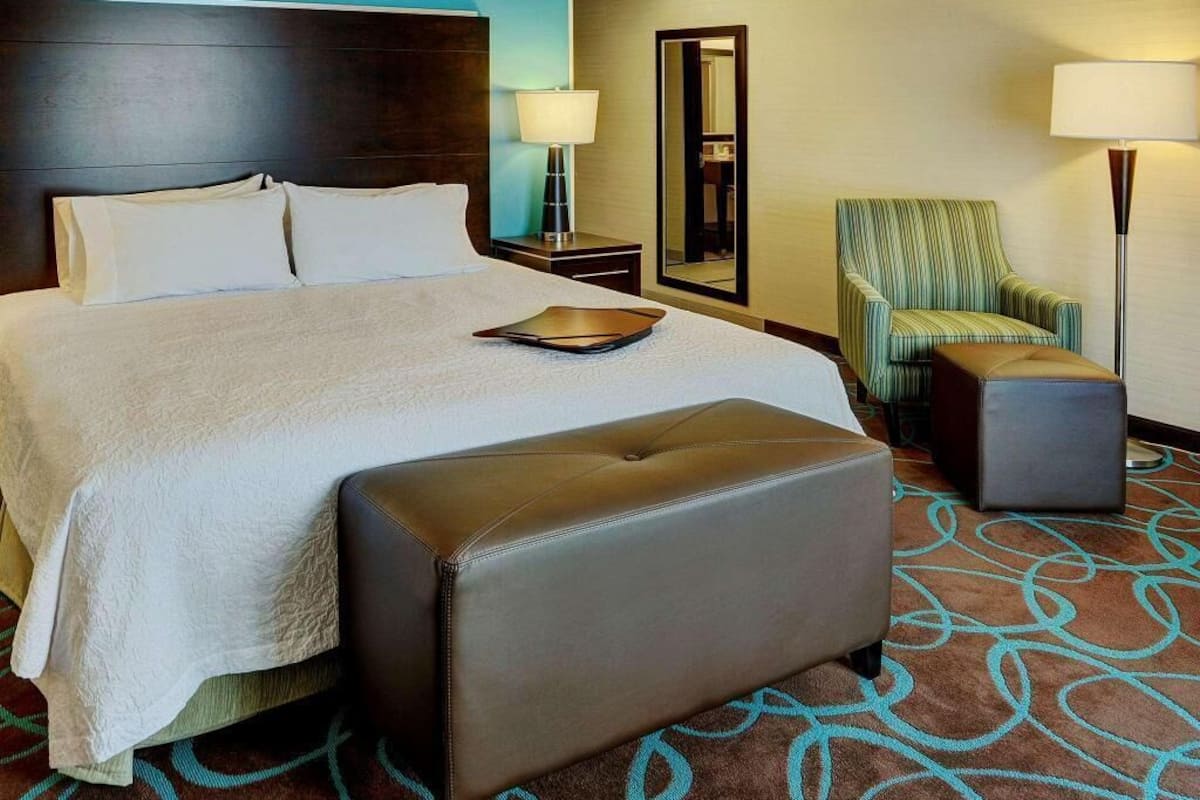 Ein Hotelzimmer mit einem großen Bett, einem Nachttisch, einem Sessel, einer Bank und einem Spiegel.