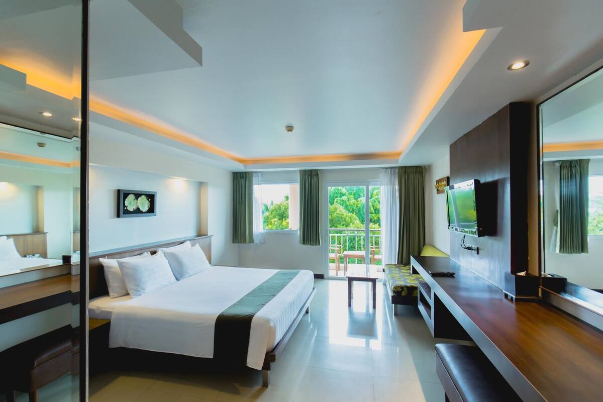 A modern hotel room with a large bed, a TV, and a balcony with a view of greenery.