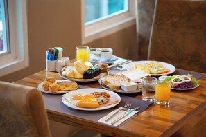 Una mesa de desayuno con platos de huevos, salchichas, tostadas y fruta. También hay tazas, un vaso de zumo de naranja y un salero y pimentero.