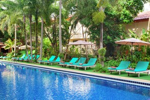 Zona de piscina con sillones de descanso y sombrillas, rodeada de palmeras y follaje.