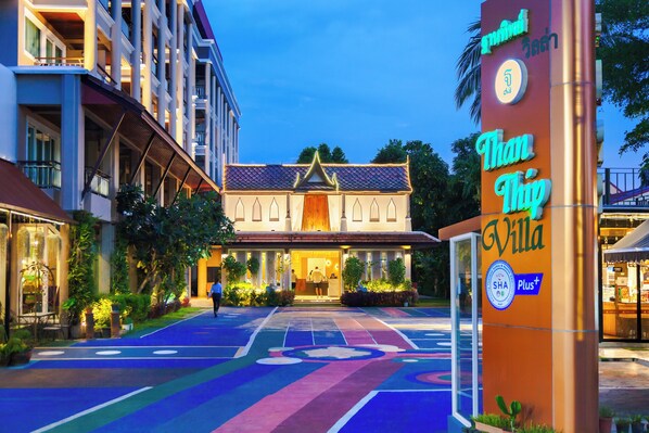 Fachada de hotel de estilo arquitectónico tailandés tradicional, con un camino colorido que conduce a la entrada, y un cartel que indica el nombre del hotel y sus servicios.