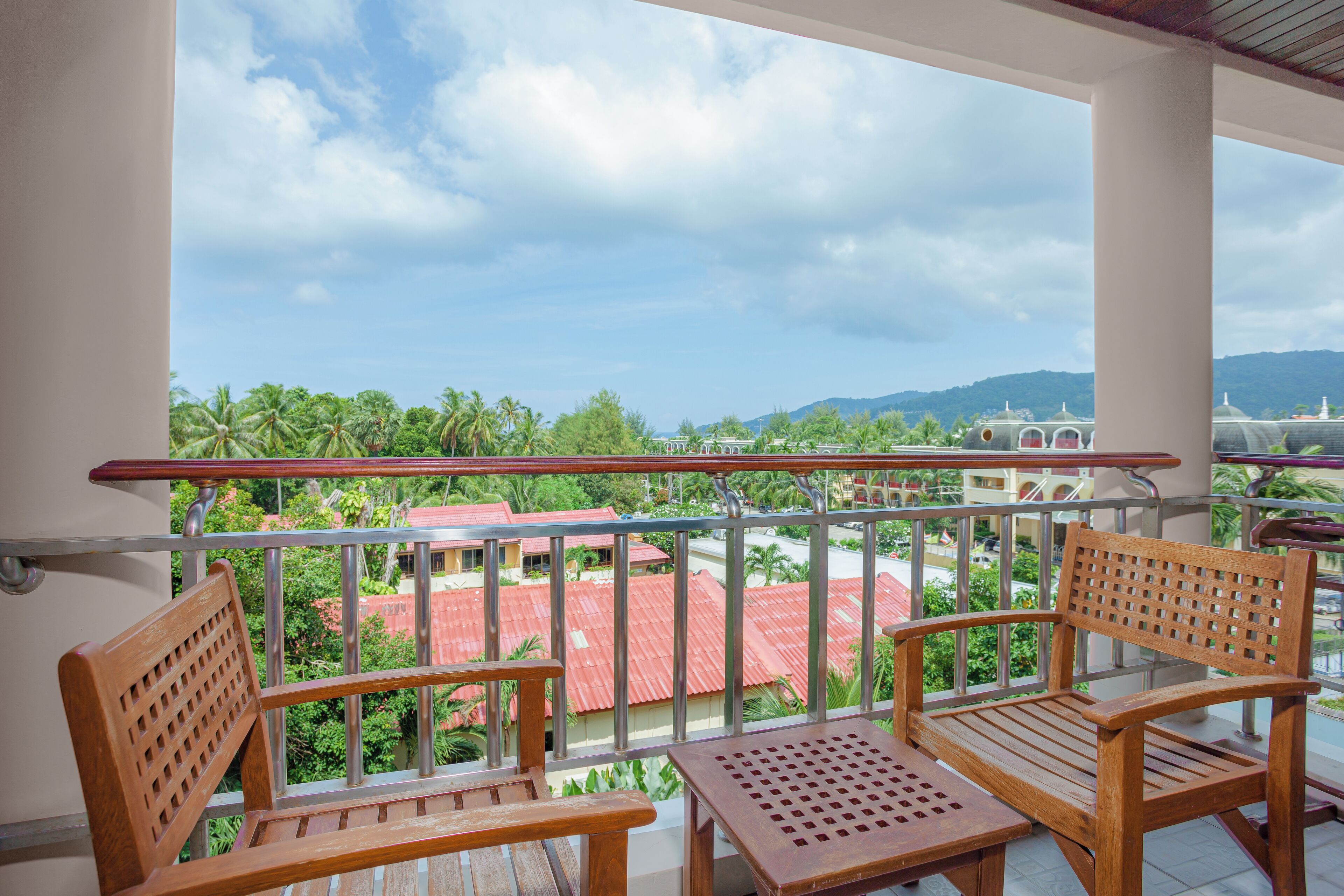A balcony with two wooden chairs and a small table, overlooking a red-roofed building and lush greenery.