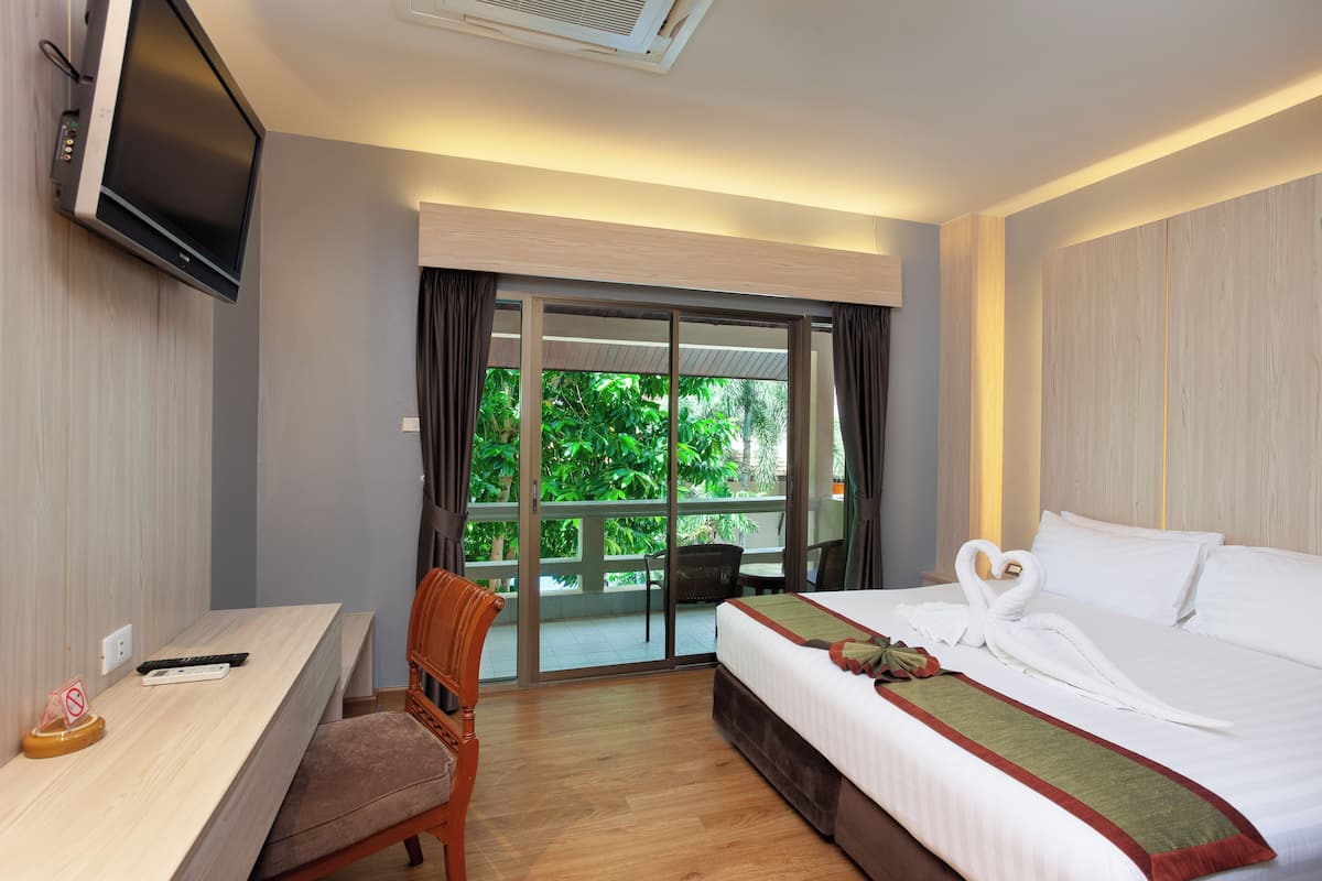 A hotel room with a bed, a desk, a chair, a TV, and a view of greenery through the window.