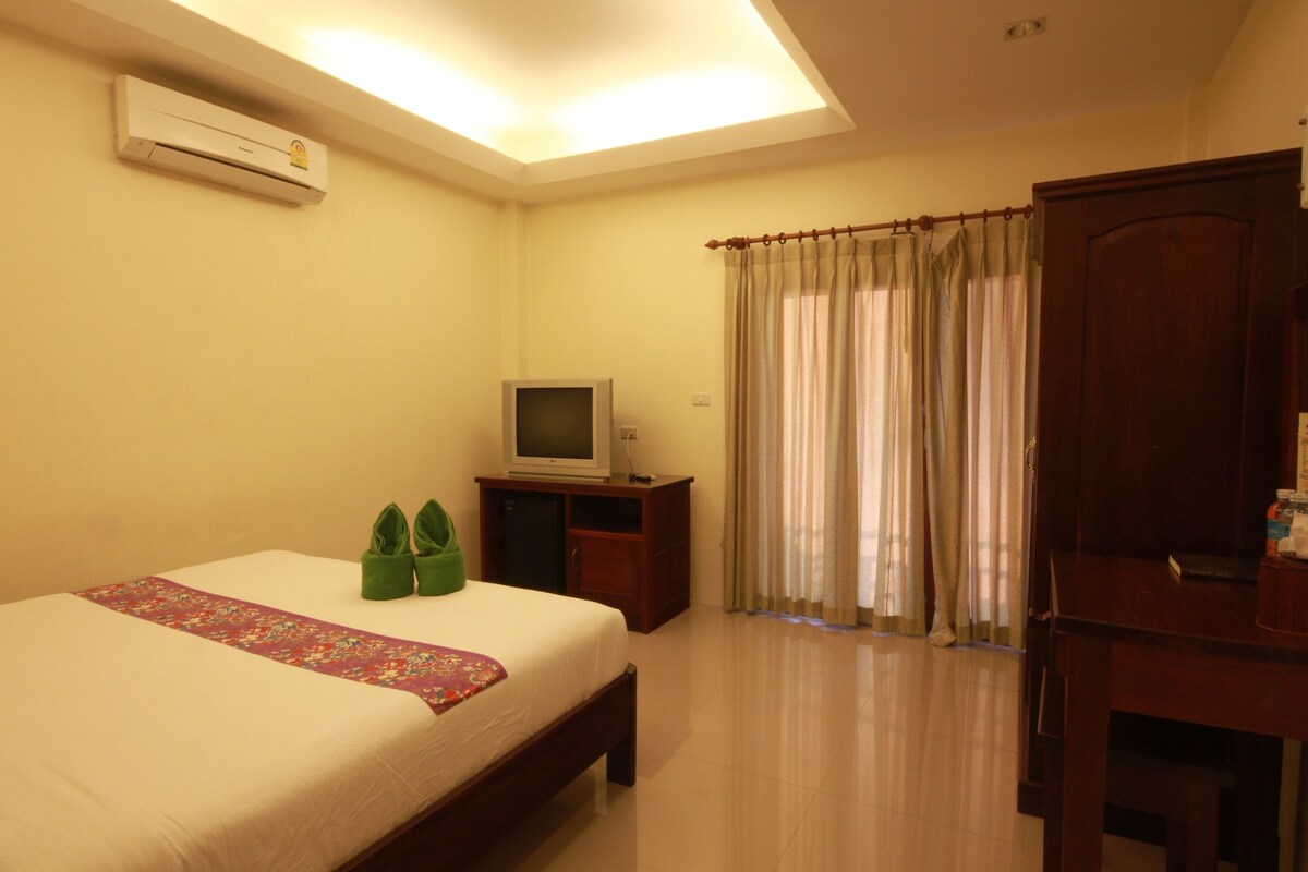 A hotel room with a bed, a television on a stand, an air conditioning unit, a wardrobe, and a window with curtains.