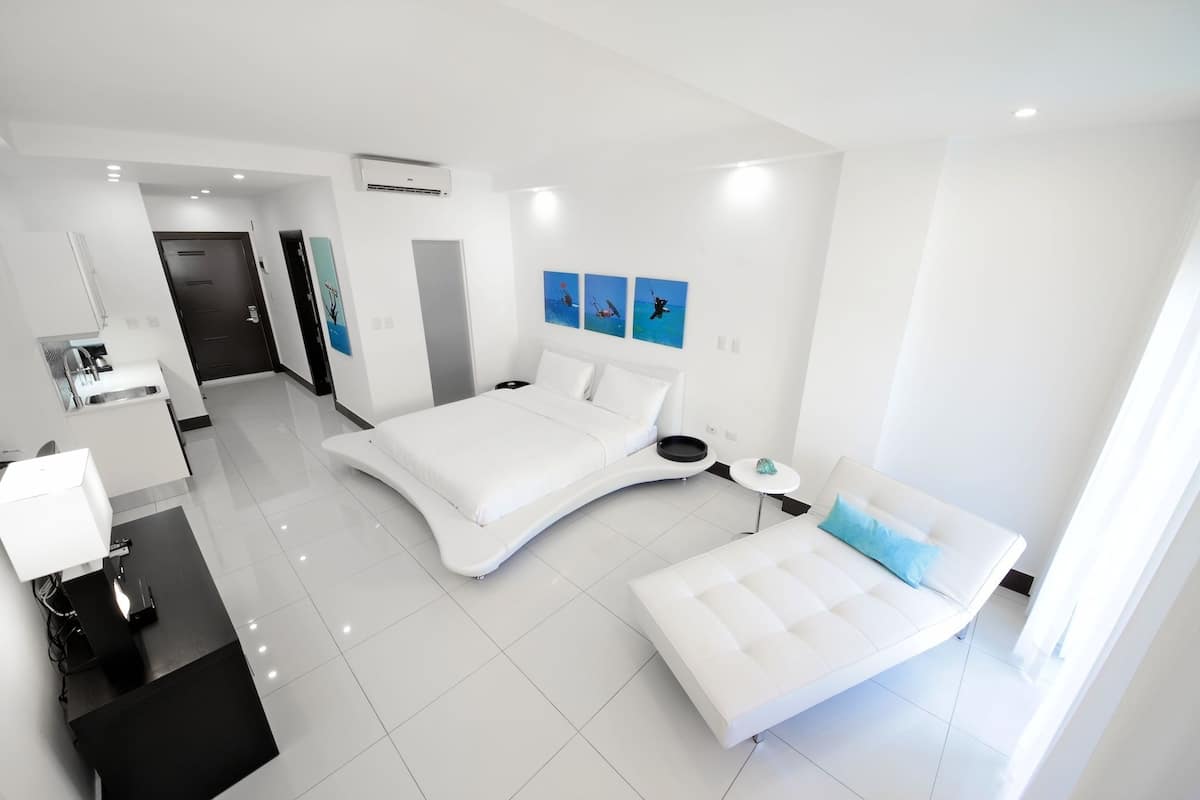 A modern hotel room with a curved bed, a bedside table, a desk with a lamp, and a view of the ocean through the window.