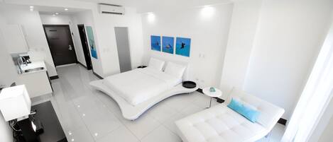 A modern hotel room with a curved bed, a bedside table, a desk with a lamp, and a view of the ocean through the window.