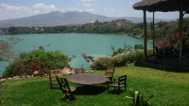 A grassy area with a round table and chairs, a thatched-roof structure, and a view of a lake with mountains in the background. - Babogaya Lake Viewpoint Lodge (Bishoftu)