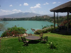 A grassy area with a round table and chairs, a thatched-roof structure, and a view of a lake with mountains in the background.