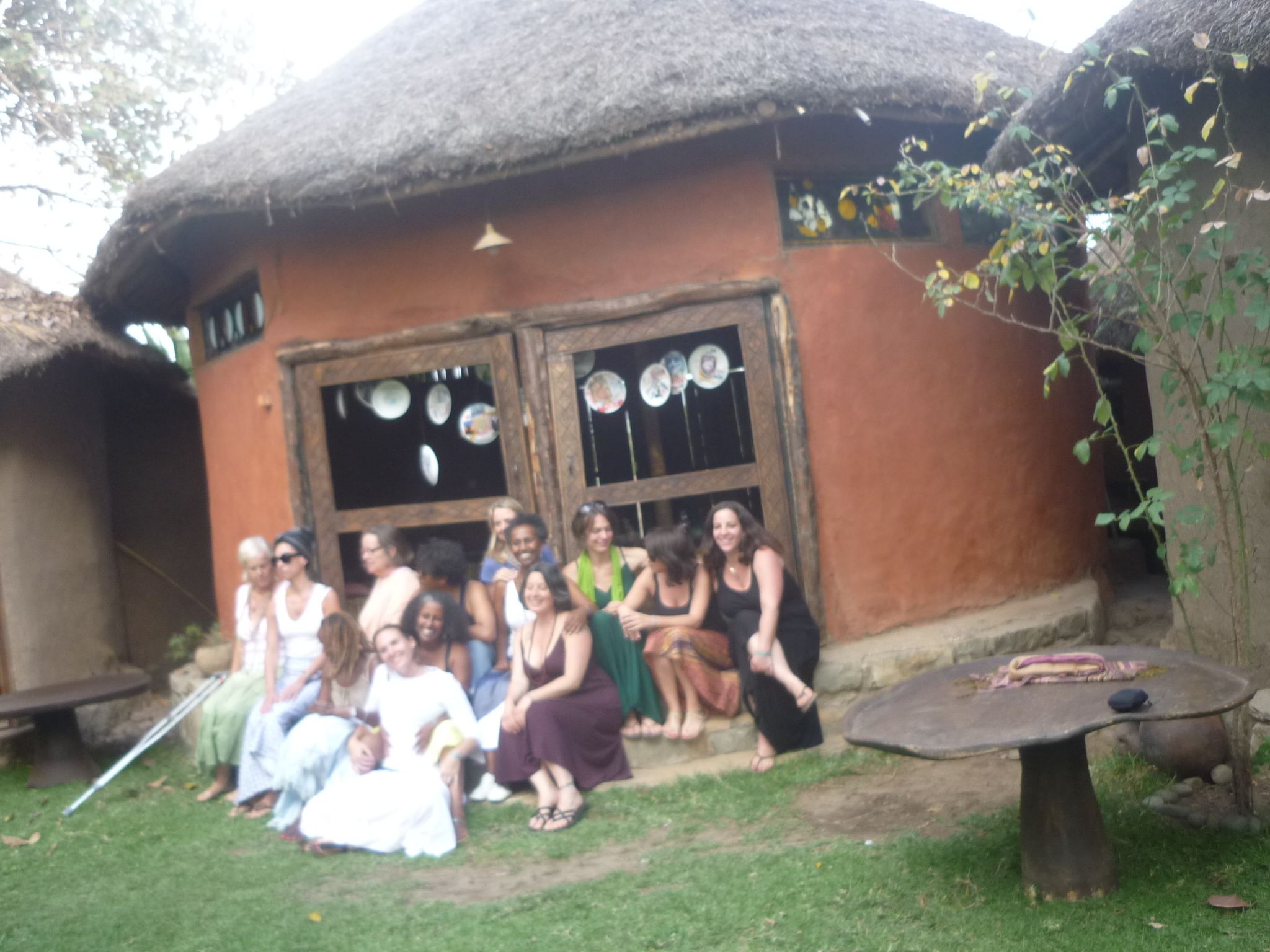 A rustic building with a thatched roof, a group of people sitting outside, and a small table with a blanket.