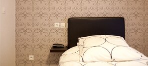 A bedroom with a patterned wallpaper, a black headboard, a bedside table with a lamp, and a bed with white bedding. - Hôtel Bel Air (Paris)
