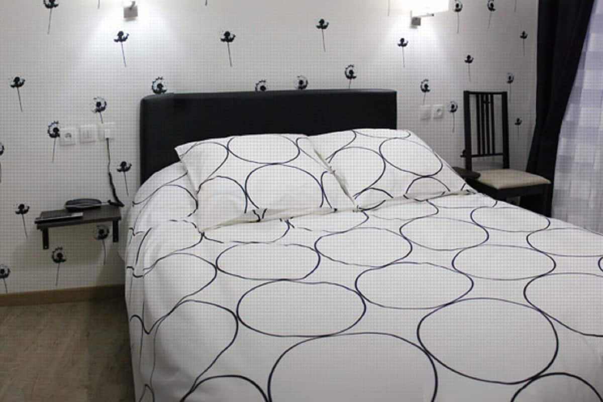 Een net opgemaakt bed met een deken met een cirkelmotief, een nachtkastje, een stoel en een wand met behang met een bloemenpatroon.