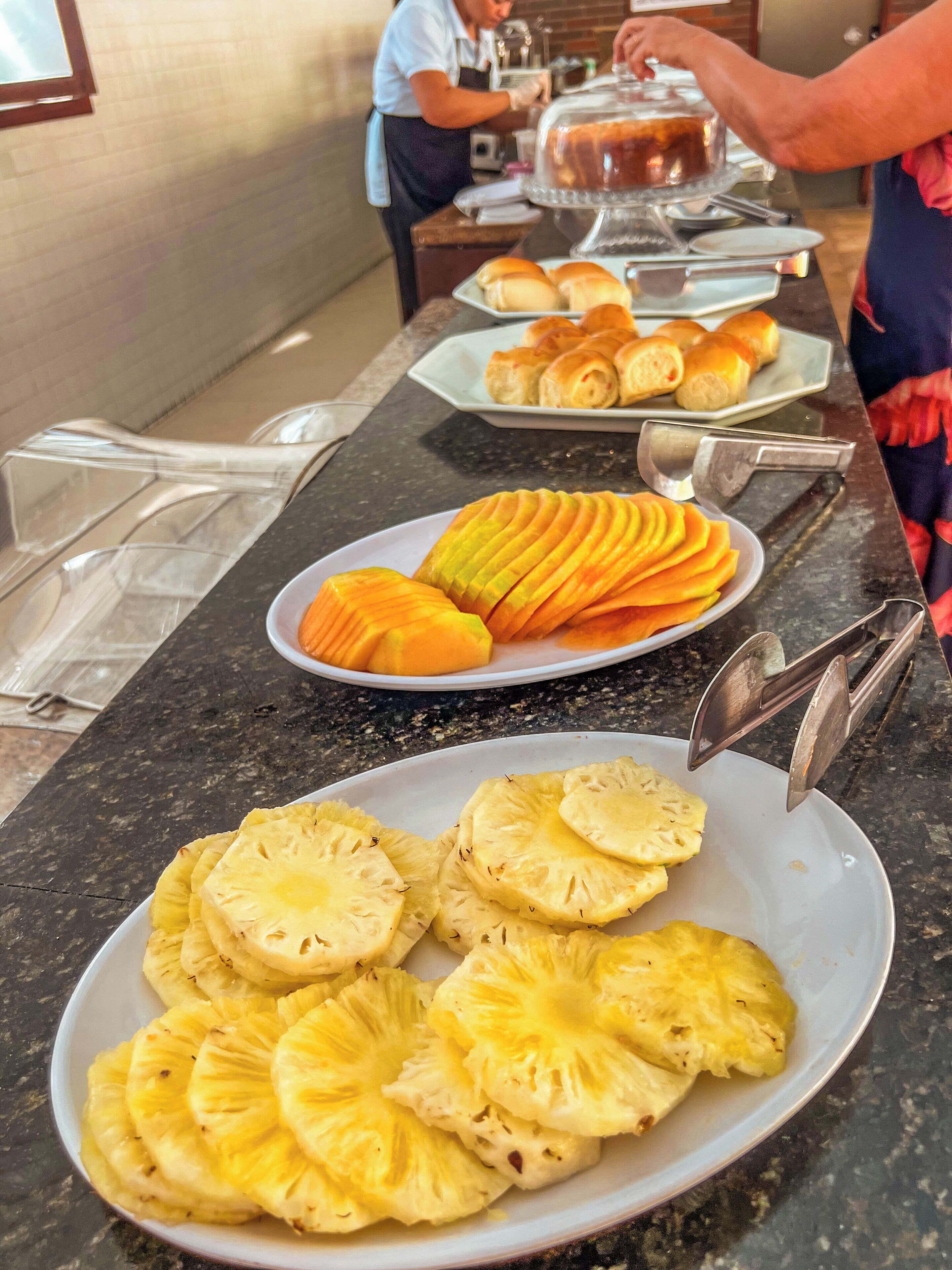 A buffet counter with plates of sliced pineapple and mango, and a person serving food in the background.