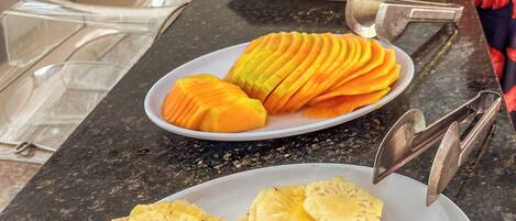 A buffet counter with plates of sliced pineapple and mango, and a person serving food in the background.