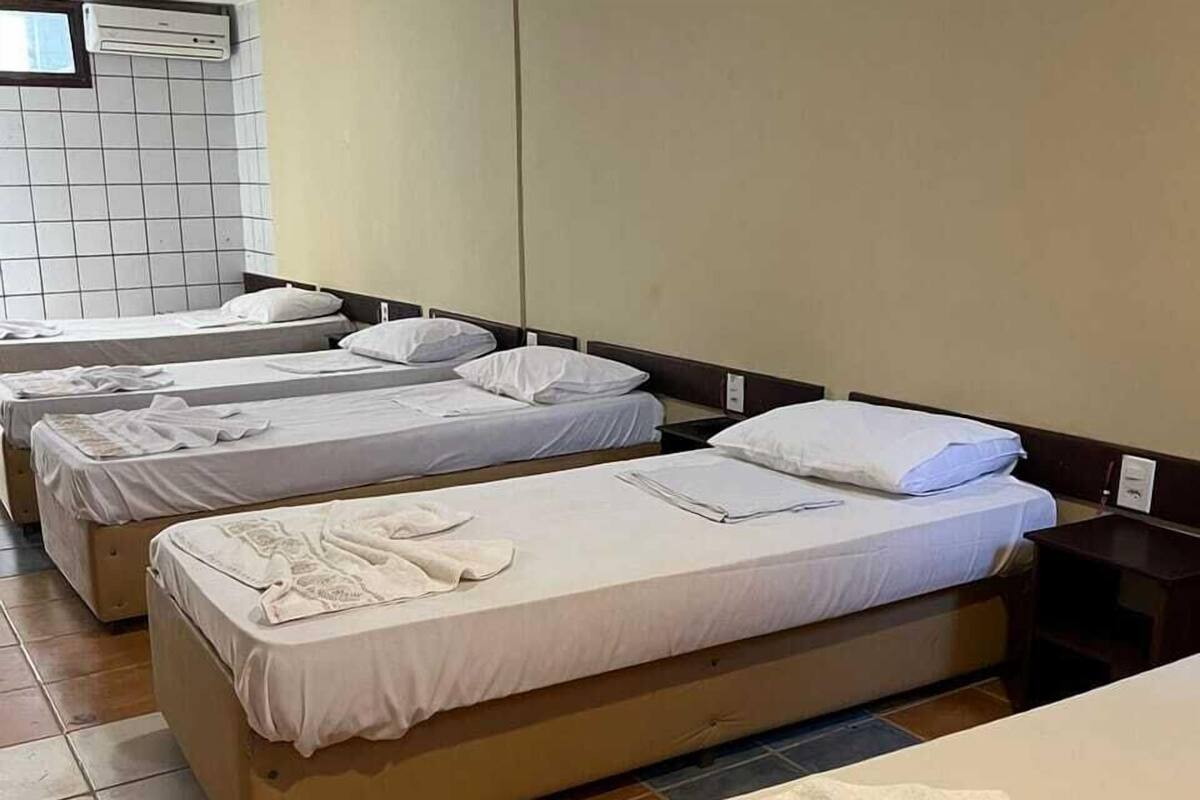 Habitació d'hotel amb tres llits individuals, cadascun amb un llençol i coixins blancs. Hi ha un panell de capçal de fusta, una taula de nit i un aire condicionat fixat a la paret.