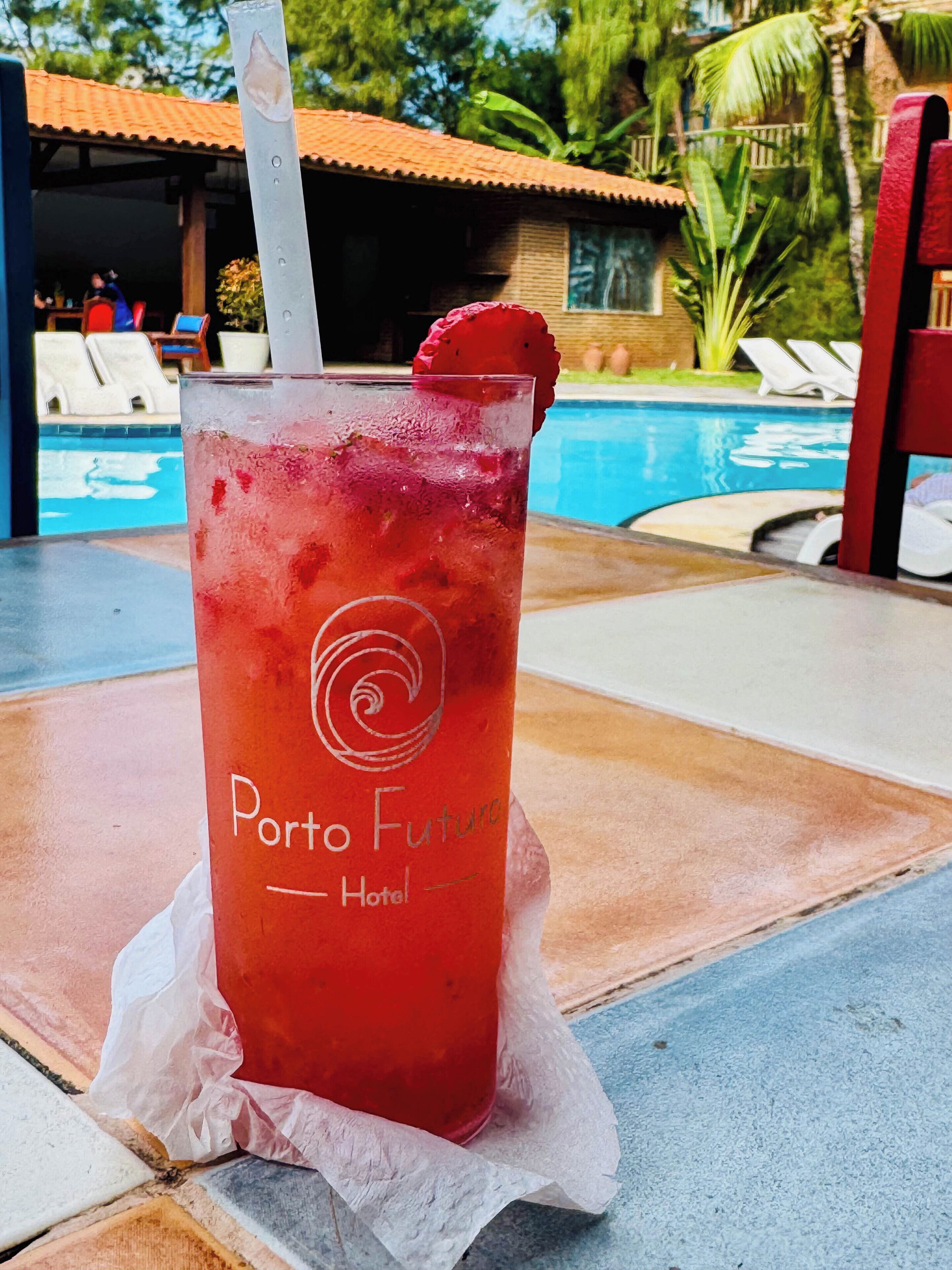 A glass of red drink with a straw and a strawberry on the rim, labeled "Porto Futuro Hotel," on a table by a poolside with lounge chairs and a palm tree in the background.