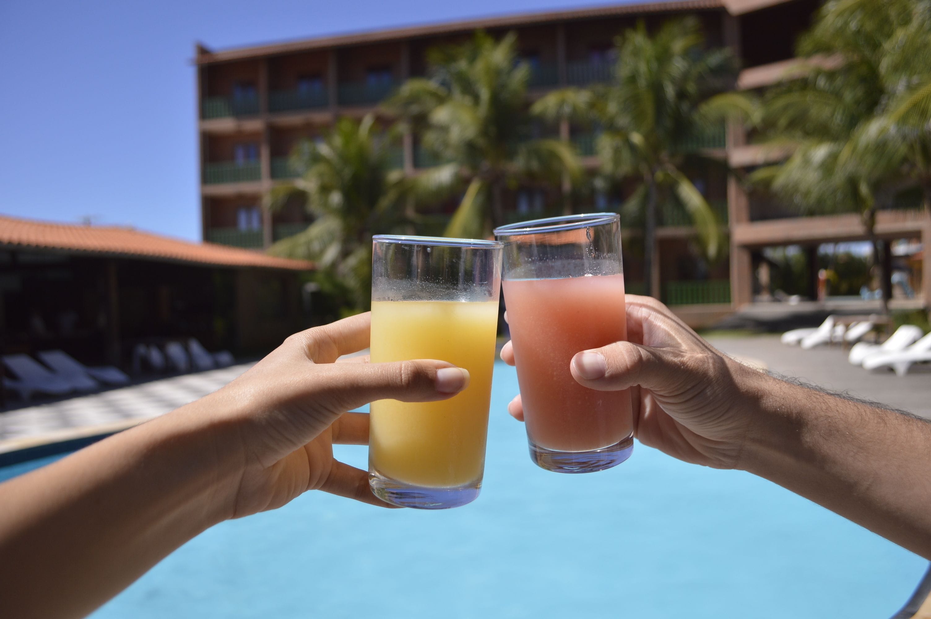 Two hands holding glasses of orange and pink beverages by a pool with a hotel building and palm trees in the background.