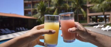Two hands holding glasses of orange and pink beverages by a pool with a hotel building and palm trees in the background.