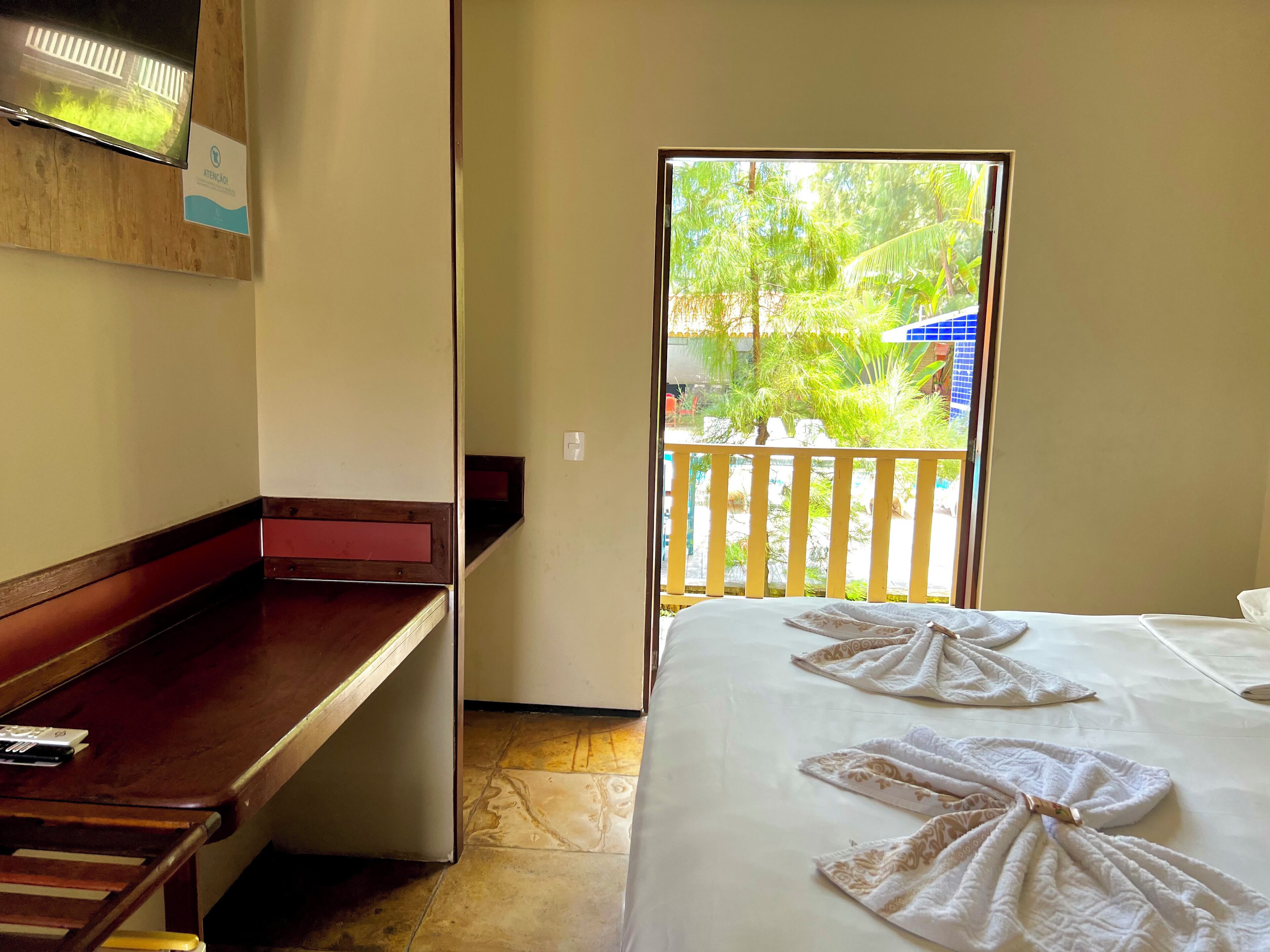 A hotel room with a bed, a wooden bench, a TV, and a balcony with a view of greenery and a building.