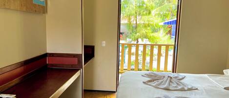 A hotel room with a bed, a wooden bench, a TV, and a balcony with a view of greenery and a building.